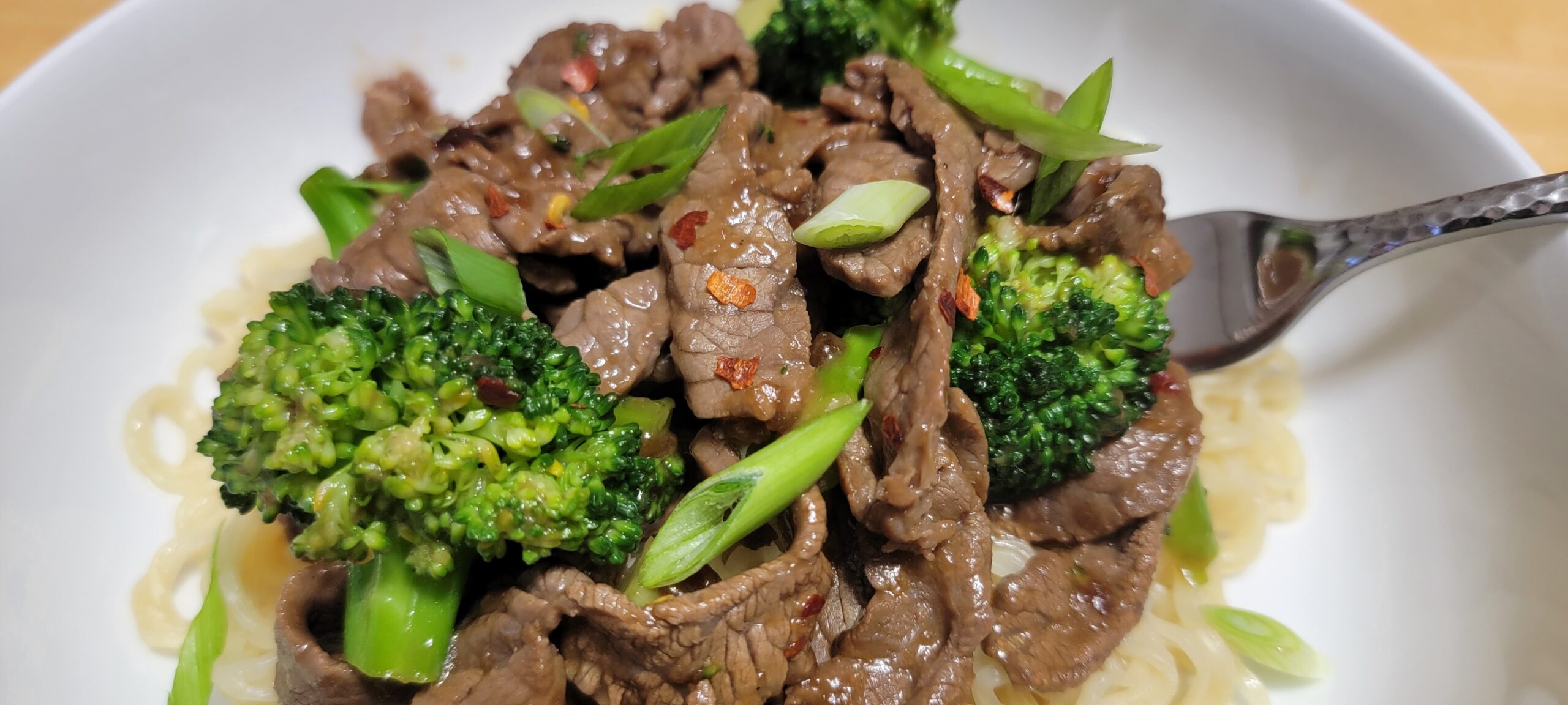 a white plate with beef and broccoli on a bed of ramen